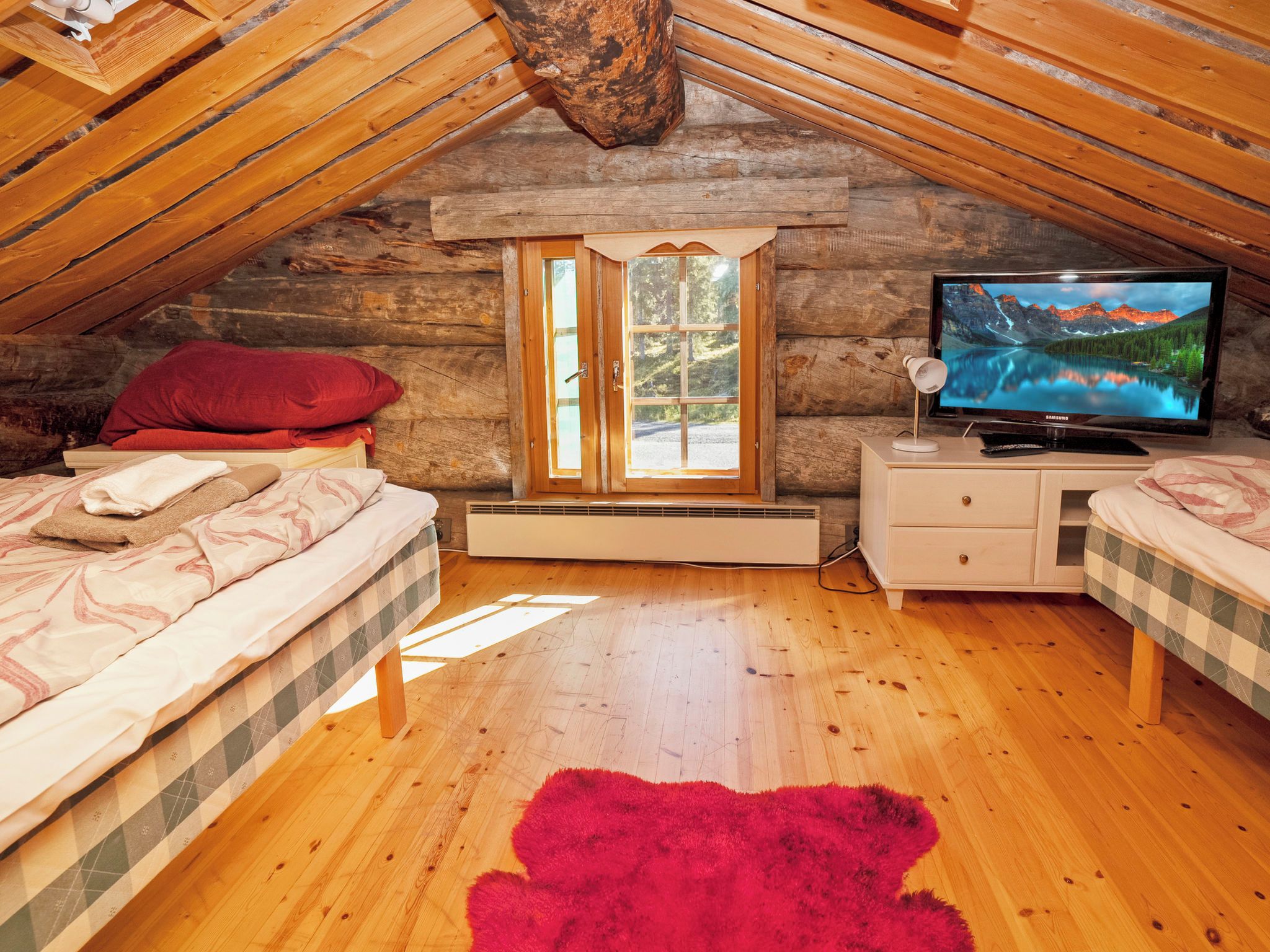 Un dormitorio acogedor en el ático con techo inclinado de madera, un ventanal grande, un televisor y dos camas con ropa de cama a cuadros.