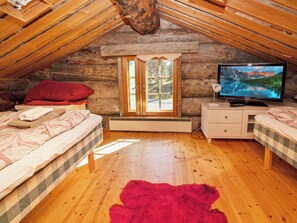 Une chambre mansardée chaleureuse avec un plafond en pente en bois, une grande fenêtre, un téléviseur et deux lits dotés de draps à carreaux.