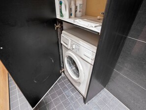 A compact laundry room with a washing machine, a shelf with toiletries, and a tiled floor. - Villa vuosselinjärvi by Interhome (Kuusamo)