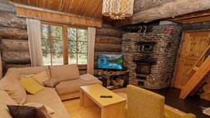 Interior de una cabaña de troncos con sofá, televisión, chimenea de piedra y mesa de comedor de madera.