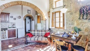 Un interior rústico con pared de piedra, vigas de madera, sofá a cuadros, una mesa de comedor con sillas y una zona de cocina.