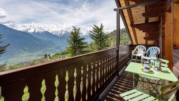 Un balcon en bois, agrémenté d’une table et de chaises, qui offre une vue sur un paysage montagneux aux sommets enneigés.