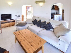Una sala de estar con un sofá, una mesa de centro de madera, un televisor sobre un mueble de madera, y una cocina visible a través de un arco.