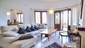 A living room with a sofa, a coffee table, and a chair. There are windows with curtains, a door, and a small table with a lamp. - Villa Clementina by Interhome (Pego)