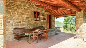 Ein Steinhaus mit einer hölzernen Bank und einem Tisch mit Stühlen, einer Holztür und Blick ins Grüne.