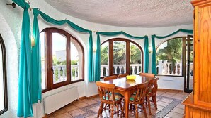 A dining area with a wooden table and chairs, large arched windows with teal curtains, and a view of greenery outside. - Vacation home Vina by Interhome (Jávea)