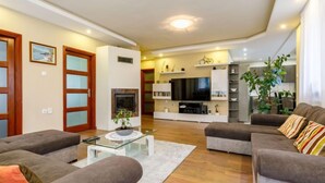 Una sala moderna con sofás grises, una mesa de centro de vidrio y una chimenea. Se ve un televisor y una estantería al fondo.