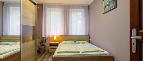 Un dormitorio con una cama de madera, una mesita de noche con lámpara, una planta en maceta y una ventana con cortinas translúcidas.