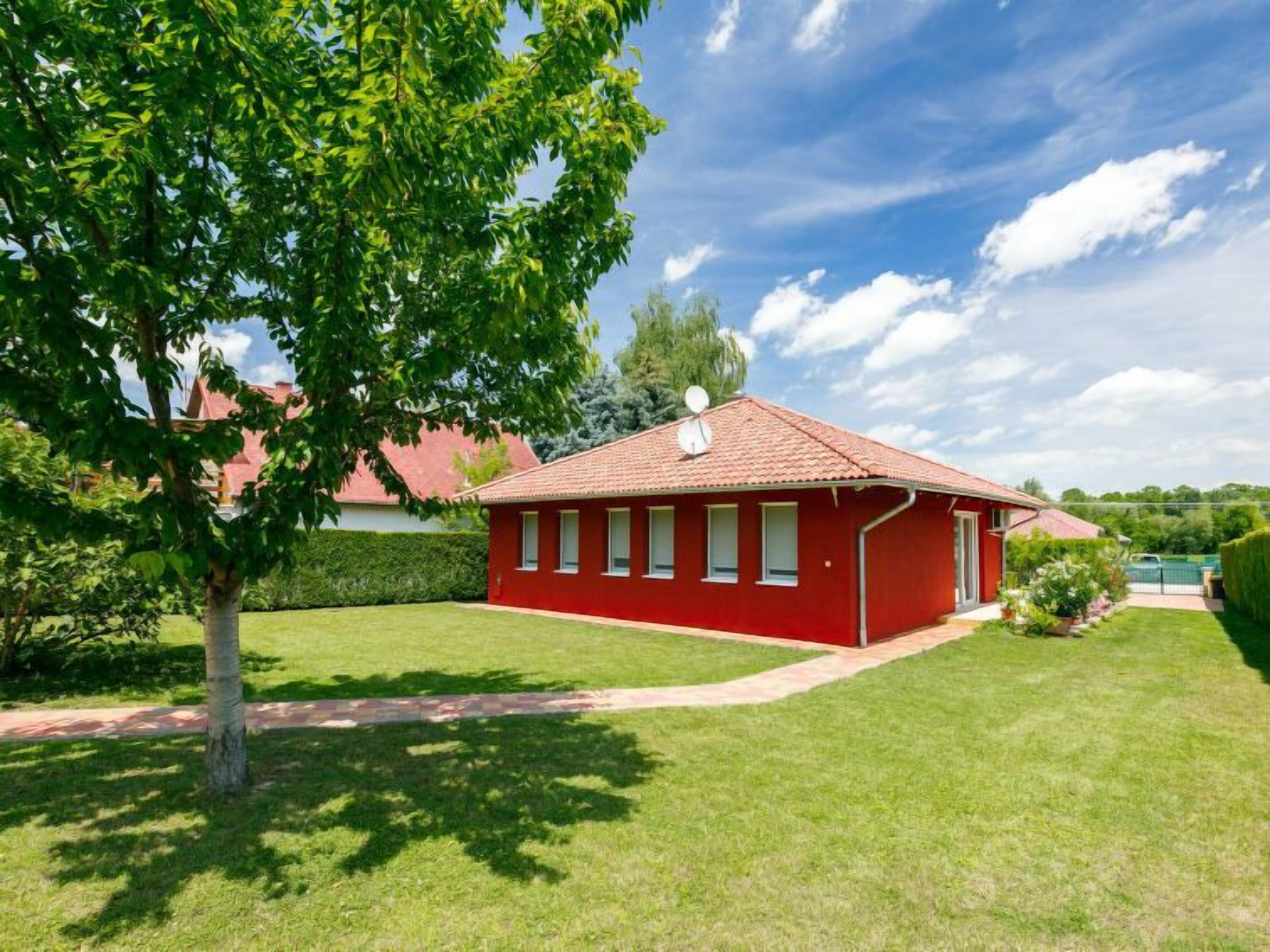 Een rode, gelijkvloerse woning met pannendak, omgeven door een verzorgd gazon en een grote boom.