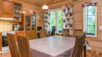 A wooden cabin interior with a dining table, chairs, a kitchen area, and a window with curtains.