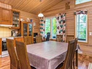 Interior de una cabaña de madera con una mesa de comedor, sillas, una zona de cocina y una ventana con cortinas.