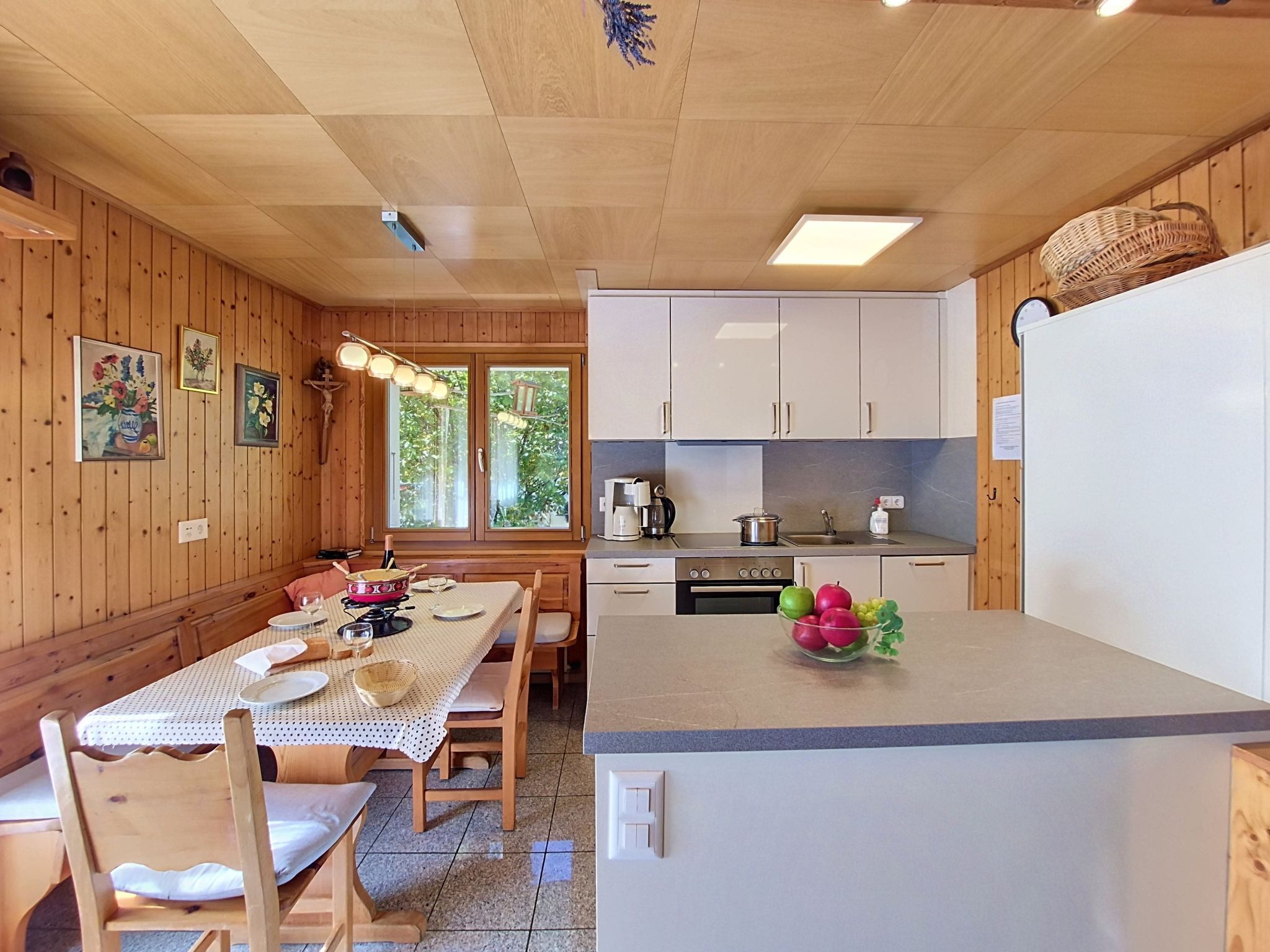 A wooden interior with a dining table, chairs, a kitchen with white cabinets, and a refrigerator.