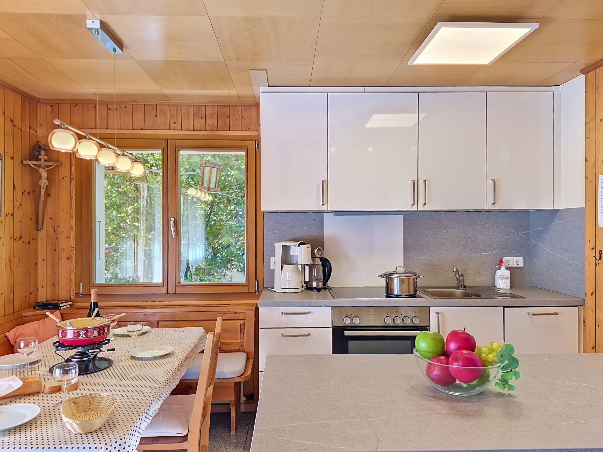 A compact kitchen with wooden cabinets, a dining table set for two, and a window with a view of greenery.