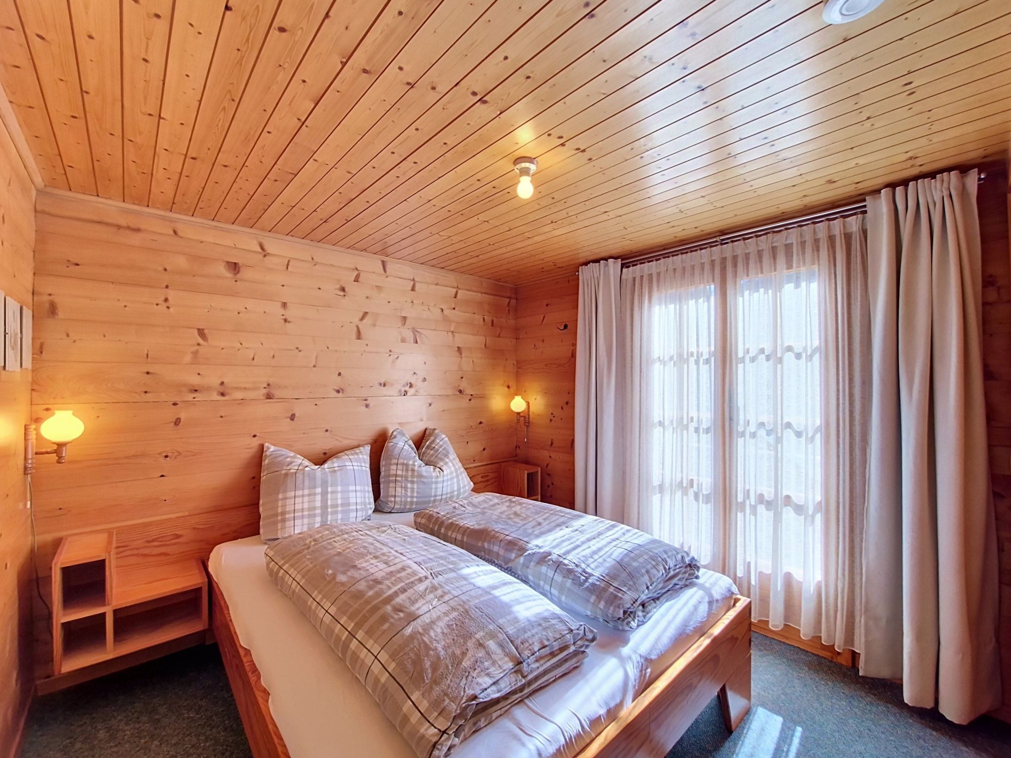 A wooden cabin bedroom with a bed, two bedside lamps, a window with curtains, and a wooden headboard.