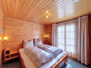 A wooden cabin bedroom with a bed, two bedside lamps, a window with curtains, and a wooden headboard. - Allegra by Interhome (St Niklaus)