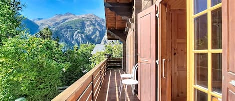 Un balcón de madera con vistas a las montañas, una puerta de madera y una pequeña mesa con sillas.