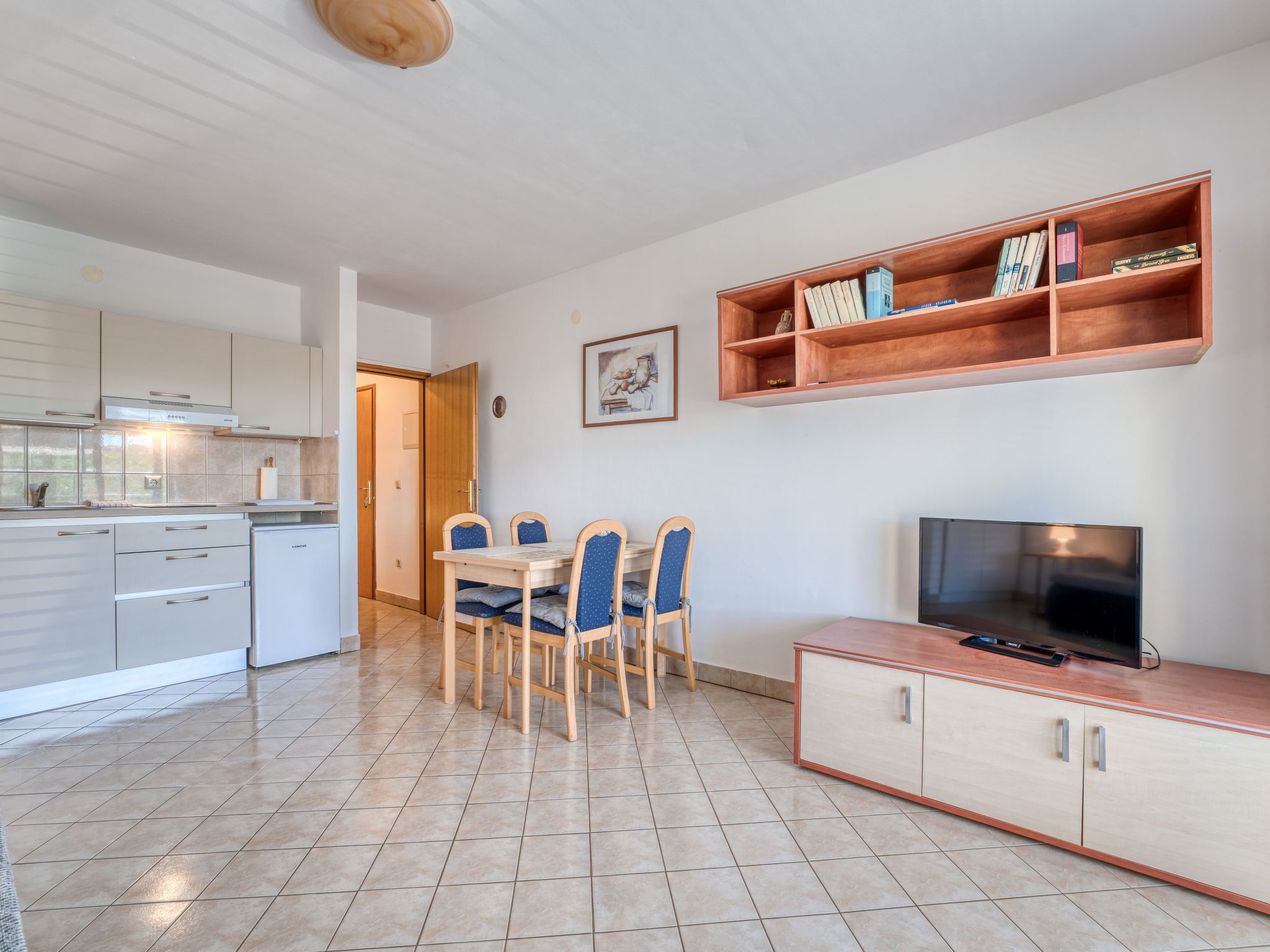 Een moderne keuken met witte kasten, een eettafel met blauwe stoelen, een televisie op een houten kast en een boekenkast met boeken.