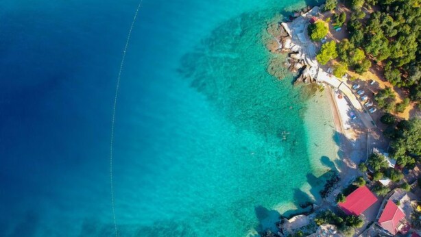 An aerial view of a coastal area with clear turquoise waters, a sandy beach, and a cluster of buildings with red roofs.
