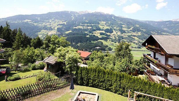 Un village rural avec des maisons traditionnelles, un jardin clos et une vue sur des collines ondoyantes et des montagnes au loin.