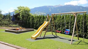 Une aire de jeux avec un toboggan et des balançoires, entourée d’une clôture et offrant une vue sur la montagne.