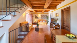Uma sala ampla com escada de madeira, um conjunto de sofás, uma mesa de jantar com uma fruteira e uma televisão.