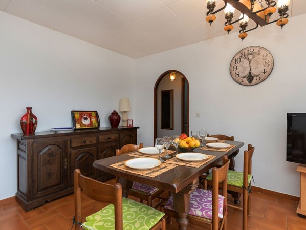 Un'area pranzo con un tavolo di legno apparecchiato per sei persone, circondato da sedie con cuscini a motivi decorativi. Si notano un orologio a parete grande e uno specchio ornamentale.