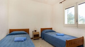 Una habitación sencilla y limpia con dos camas de madera, una mesita de noche con lámpara y una ventana con vistas a árboles.