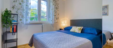 A bedroom with a bed, a nightstand, a bookshelf, a window with curtains, and a radiator.