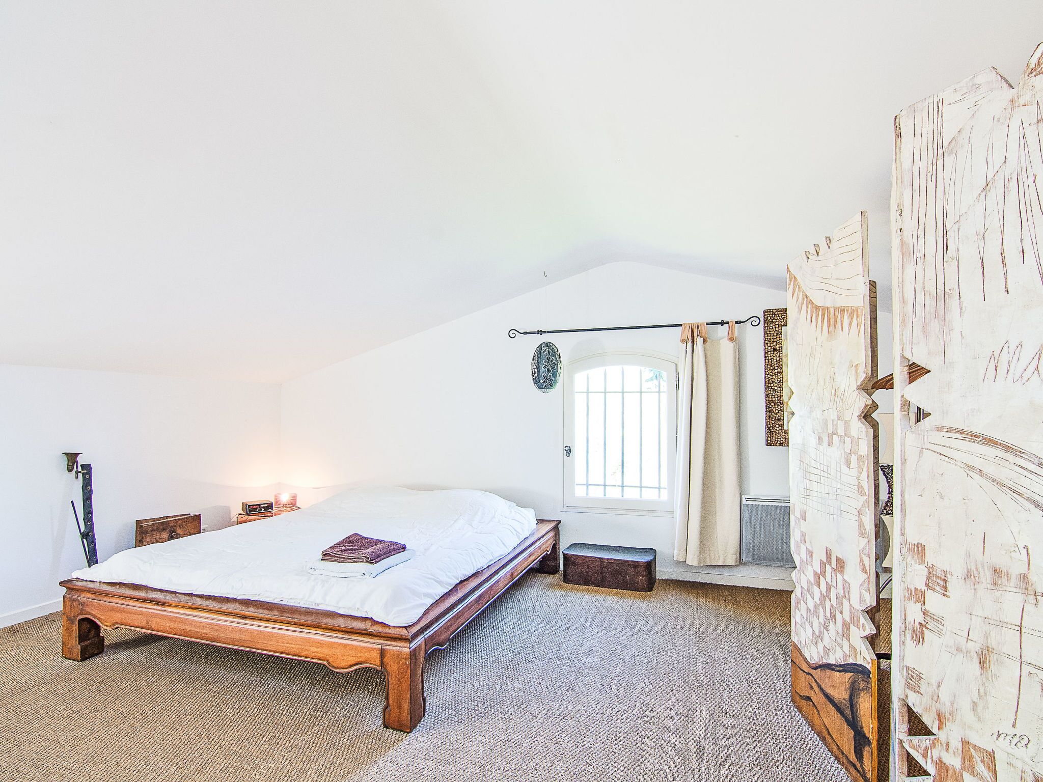 A bedroom with a wooden bed, a small bedside table, a nightstand, a window with a curtain, and a door with a decorative handle.