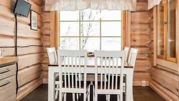 A wooden cabin interior with a dining table and chairs, a television mounted on the wall, and a window with a view of trees.