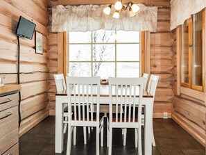 Ein Holzblockhaus-Interieur mit Esstisch und Stühlen, einem an der Wand befestigten Fernseher und einem Fenster mit Blick auf Bäume.