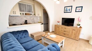 A modern living room with a blue sofa, wooden dining table, and a kitchen area with a TV and wooden cabinets. - Antoine by Interhome (Marina)