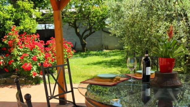 Une table ronde en verre, agrémentée d’une bouteille de vin et d’un vase rouge, est placée à l’extérieur, avec une chaise et un chemin de table.