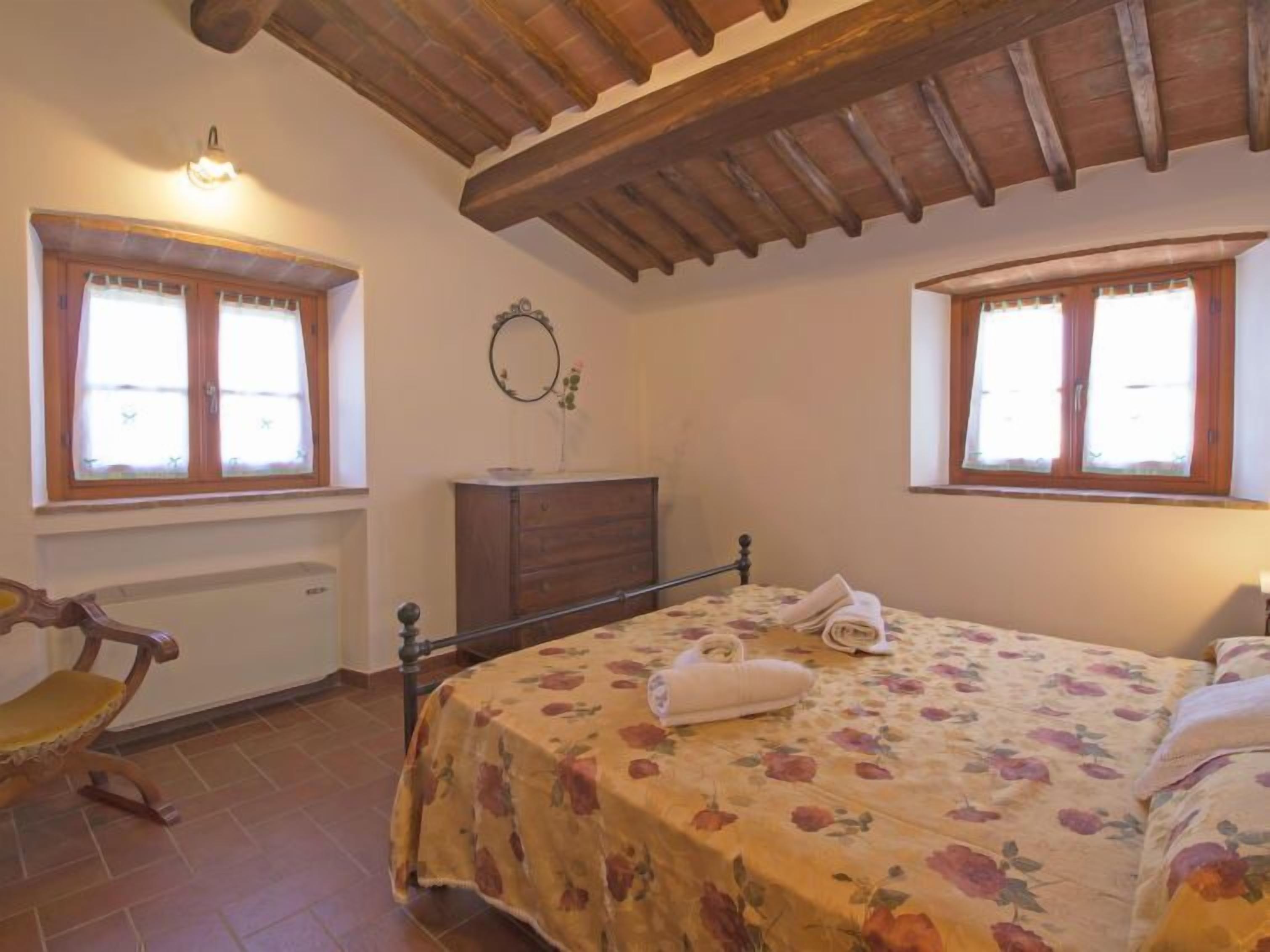 Quarto com teto de madeira, duas janelas com cortinas, uma cama com roupa de cama floral, uma mesa de cabeceira e um aparador.