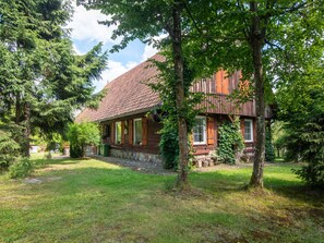 Une maison en bois avec un toit en tuiles, entourée d’arbres et bordée d’une cour gazonnée.