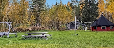 Area picnic con panchine, un piccolo deposito e un edificio rosso in un prato erboso con alberi sullo sfondo.