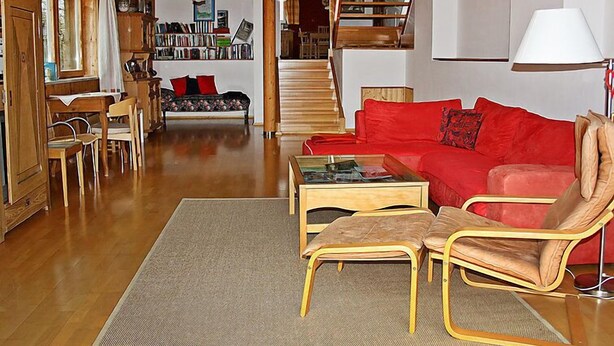 Una sala de estar acogedora con un sofá rojo, una mesa de centro de madera y dos sillones. Una escalera conduce a una zona superior con estanterías.