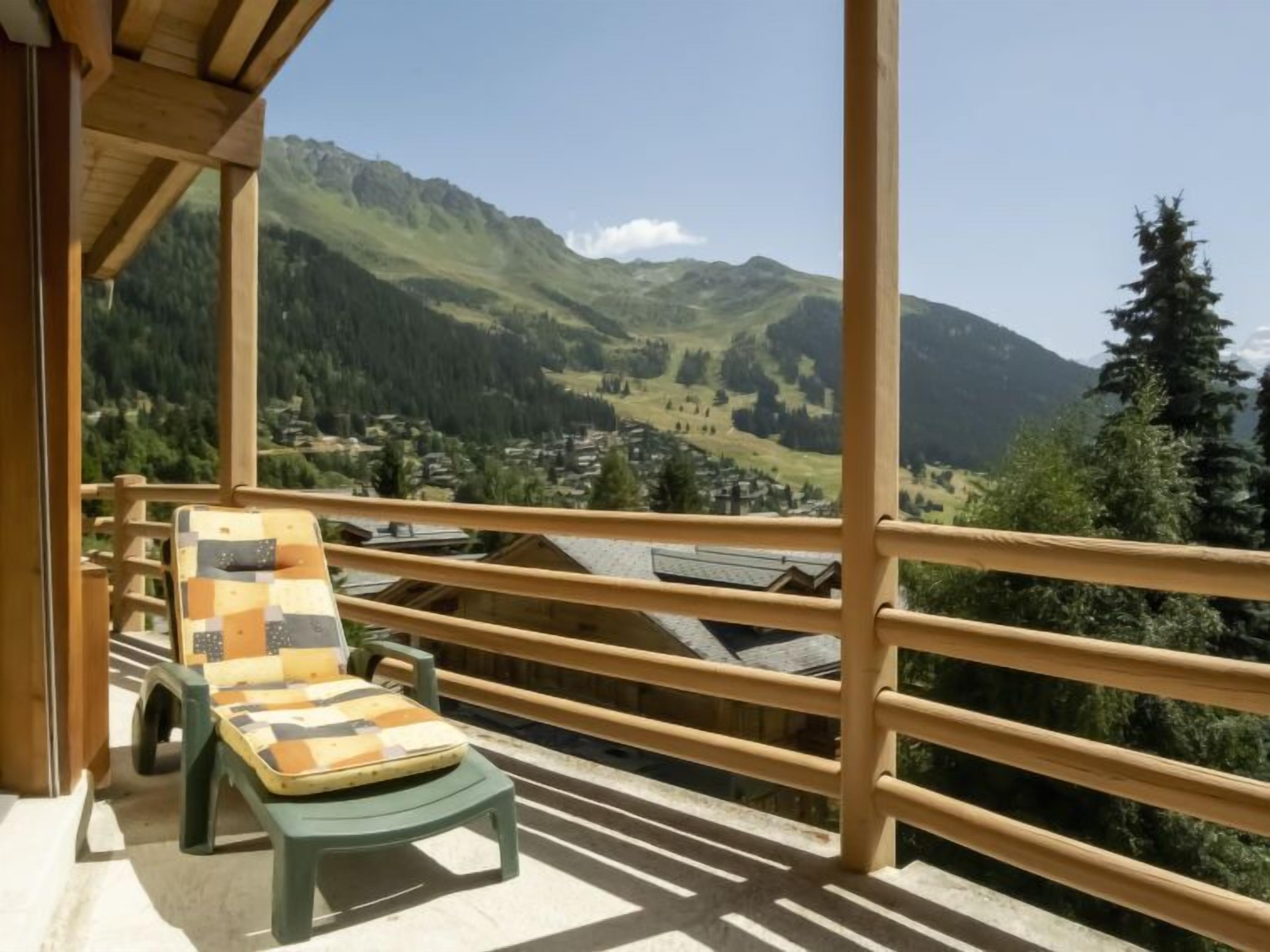 A balcony with a wooden railing, a green chair, and a cushion, overlooking a mountainous landscape with houses and trees.