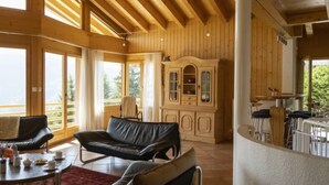 Una habitació àmplia amb un sostre alt, bigues de fusta, una finestra gran, un sofà de cuir negre, un armari de fusta i una zona de menjador amb caires.