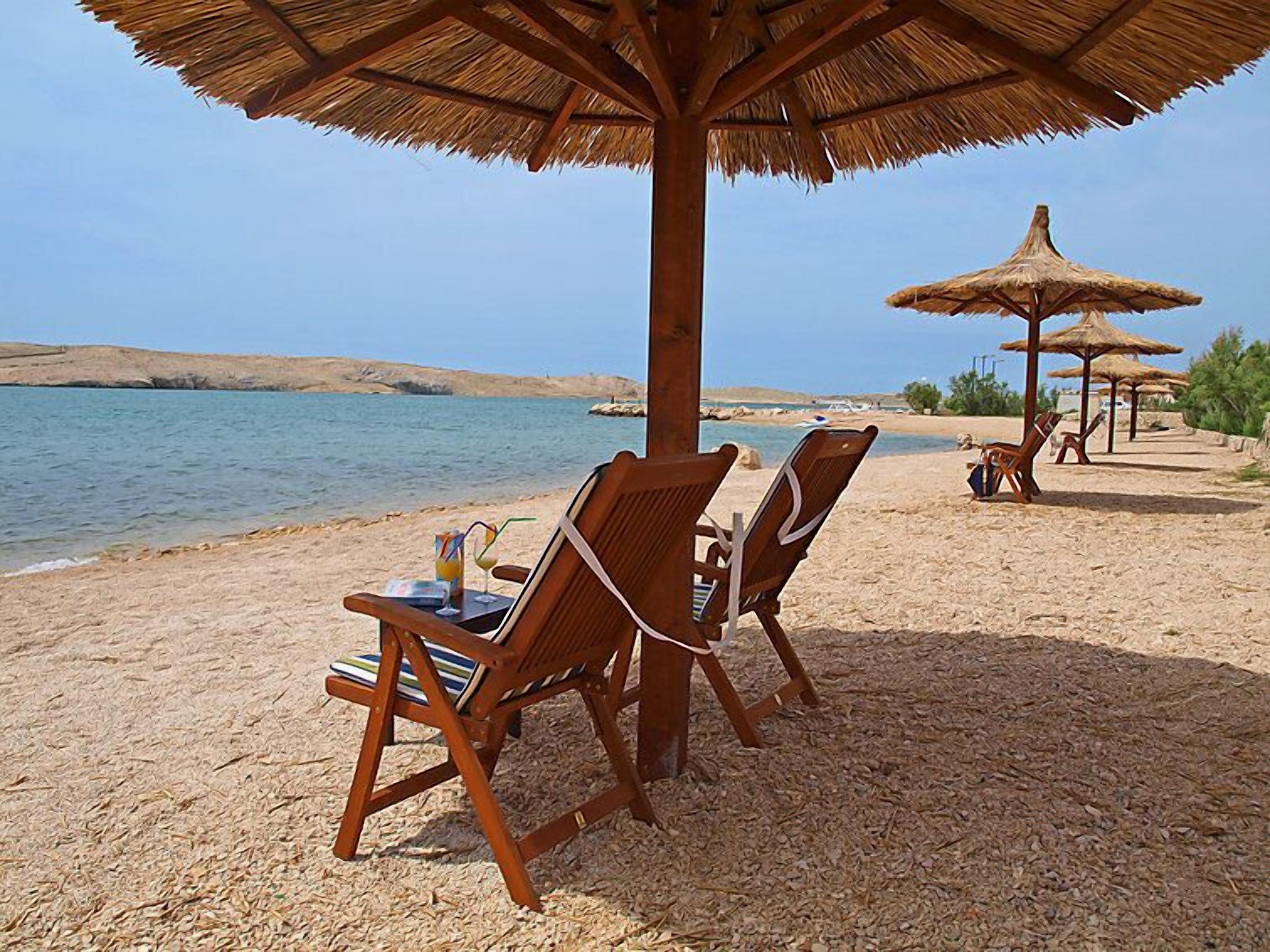 A beach with wooden lounge chairs under thatched umbrellas, a clear view of the sea, and a distant rocky shoreline.