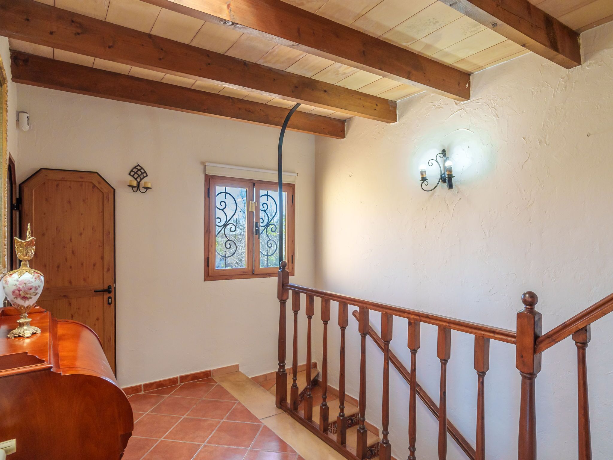 Una escalera de madera con barandilla, una puerta con pomo decorativo y un ventanal con herrajes ornamentales.