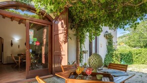 Un área de comedor al aire libre con mesa y sillas de madera, rodeada de vegetación y un ventanal que da a un espacio interior.