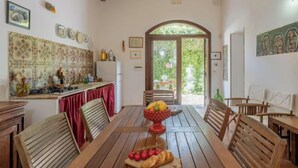 Un comedor con una mesa de madera, sillas y un frutero. Se observa una zona de cocina con salpicadero de azulejos y un refrigerador. La habitación tiene techo de madera y una puerta que da a un espacio exterior con vegetación.