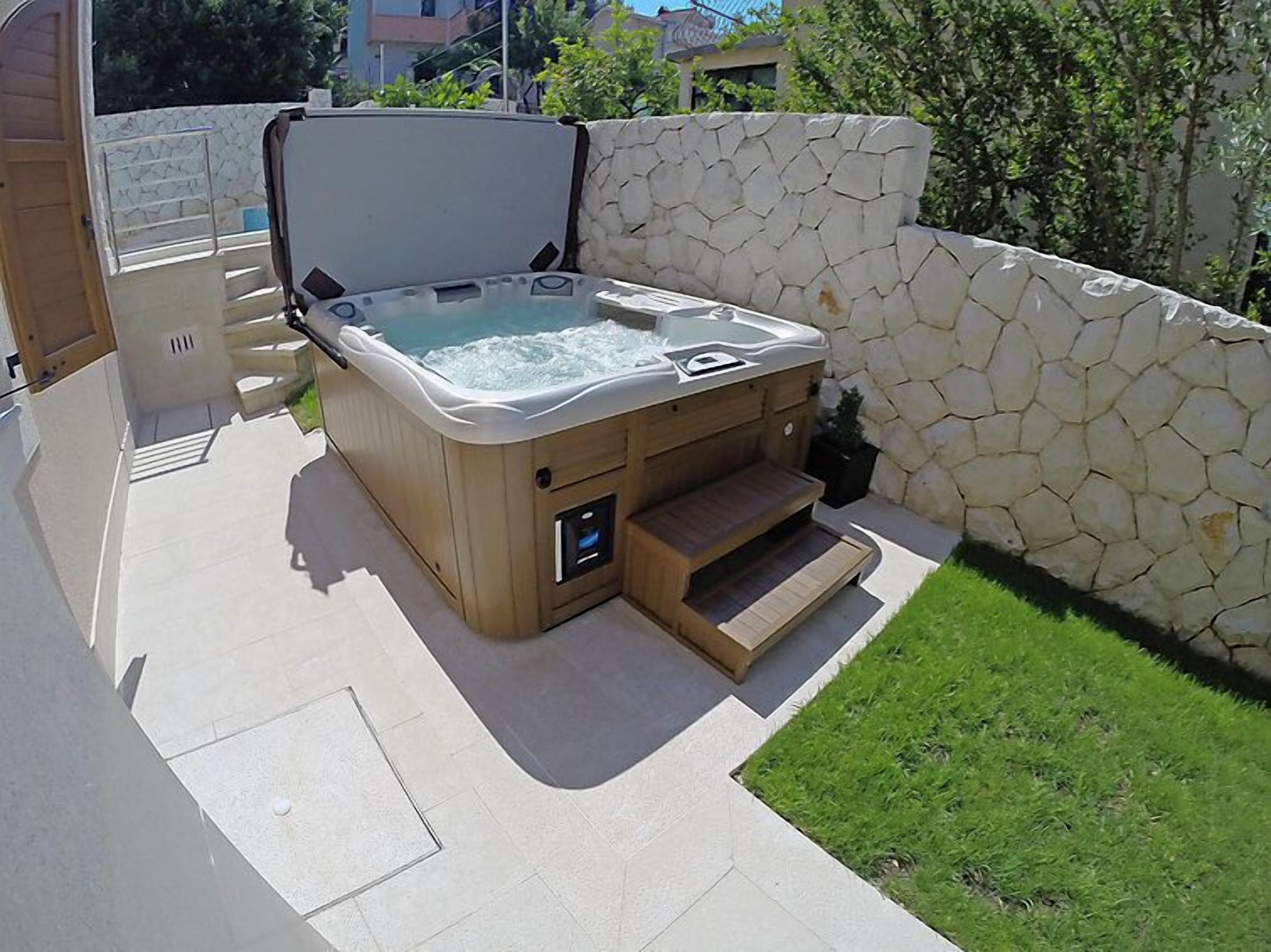 A hot tub with a wooden exterior, a control panel, and steps leading into it, set against a stone wall and a grassy area.