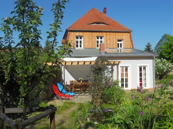 A two-story wooden house with a red roof, a covered patio with outdoor furniture, and a garden with various plants and flowers. - Gärtnerhaus by Interhome (Wurzen)