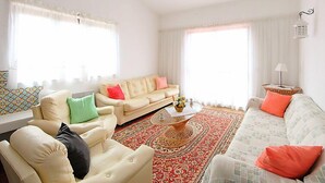 Ein Wohnzimmer mit einem beigen Sofa, einem gemusterten Sessel und einem Couchtisch. Zwei Fenster mit durchsichtigen Vorhängen, ein roter Teppich mit Muster und ein kleiner Beistelltisch mit einer Lampe.