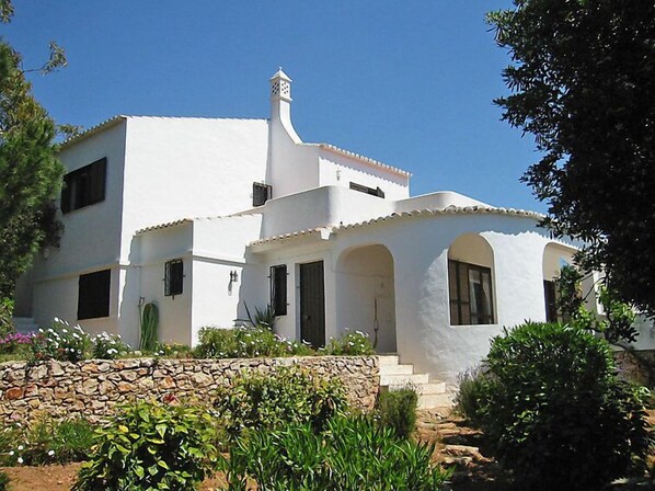 A white building with a stone wall in front, a tiled roof, and arched windows.