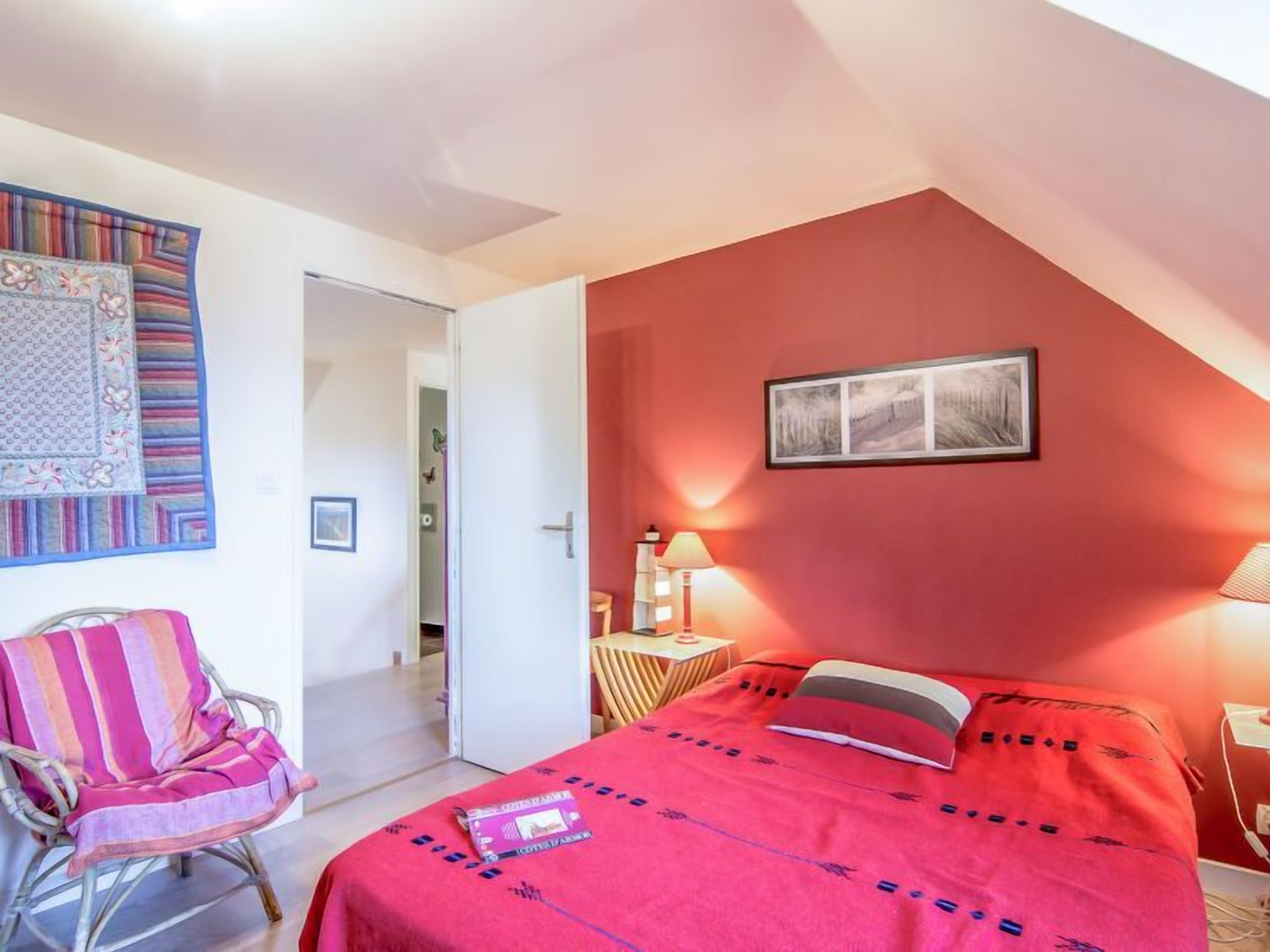 Ein Schlafzimmer mit roter Wand, einem Bett mit roter Bettdecke, einem Sessel, einem Nachttisch mit Lampe und einem gerahmten Bild an der Wand.