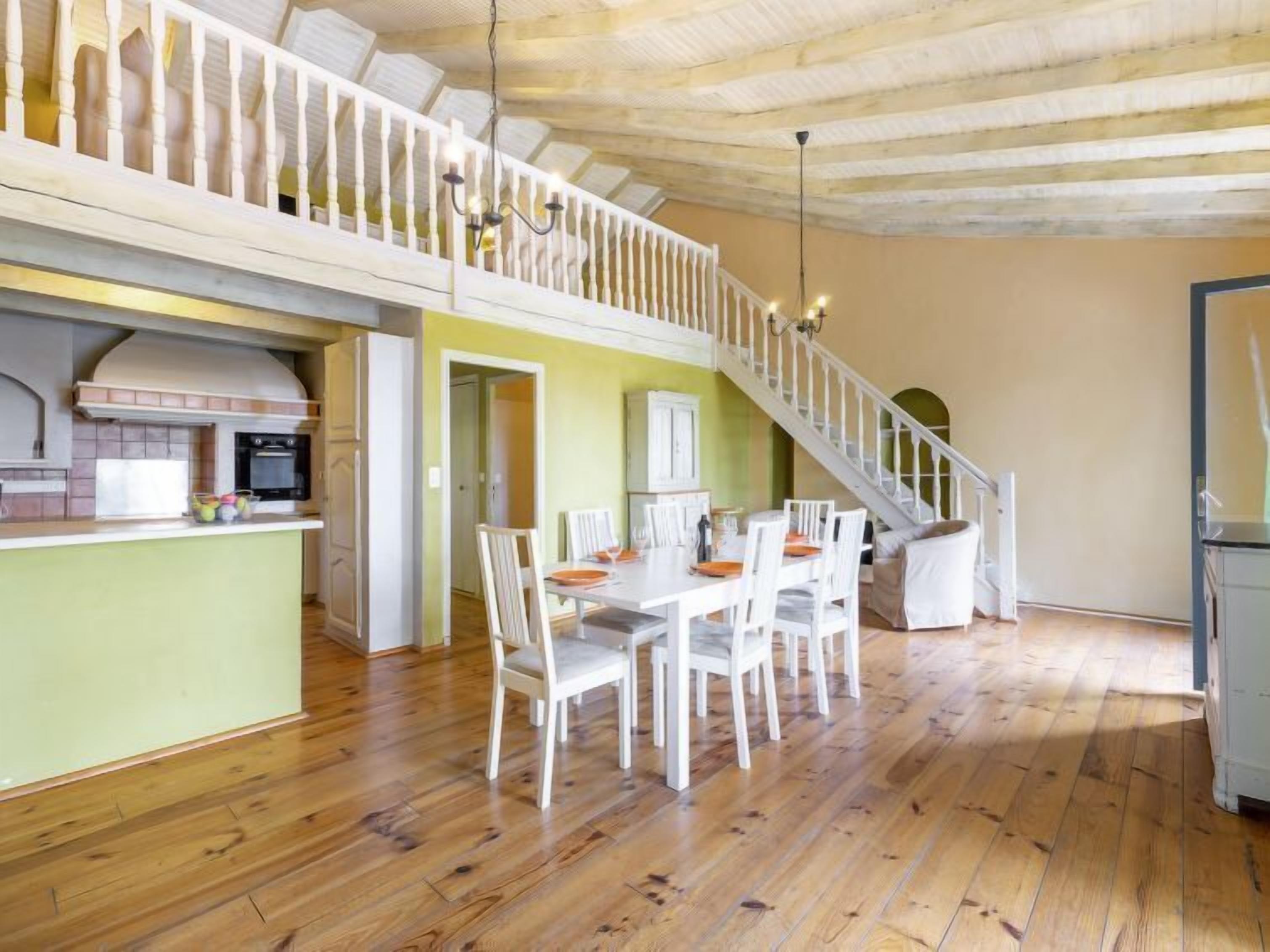 A spacious dining area with a wooden floor, white chairs, and a central table. A staircase with a white railing leads to an upper level.