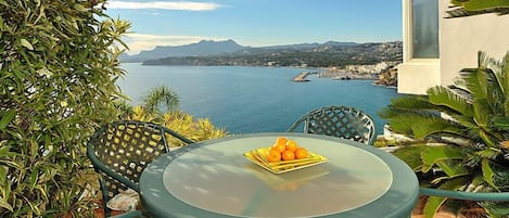 Une table ronde en verre, surmontée d’un saladier de fruits, entourée de chaises dotées de coussins à motifs floraux, et offrant une vue sur la côte, avec des montagnes et une étendue d’eau.